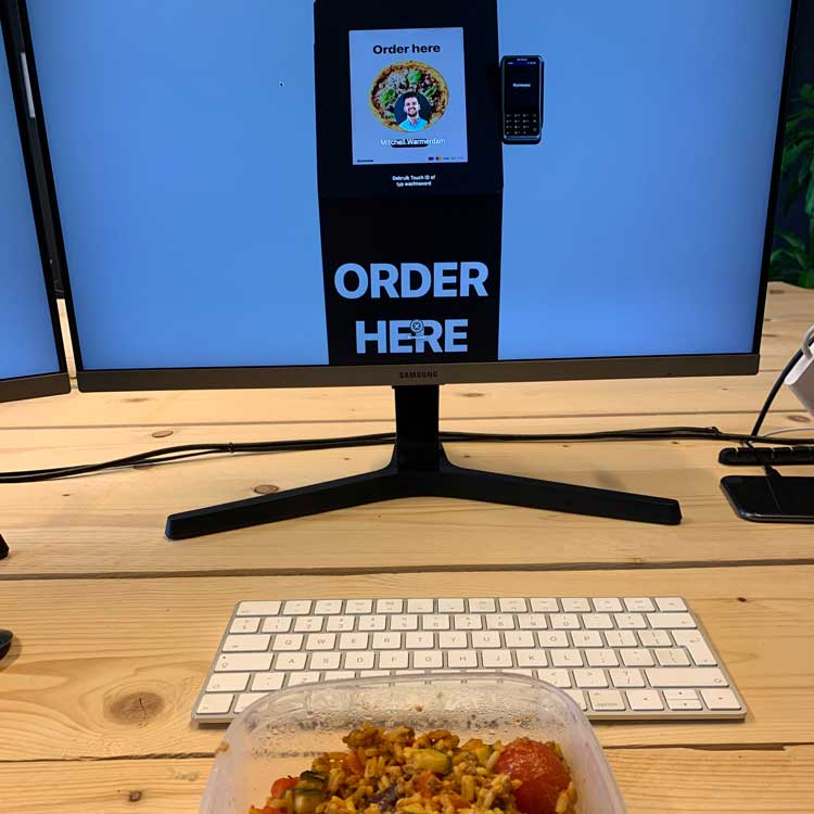 Onesix bestelzuil prototype met Order Here scherm op een bureau naast een Samsung monitor en pinautomaat.