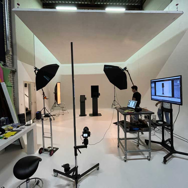 Photography studio setup with lighting equipment, cameras on stands, a white backdrop, and a person working at a laptop connected to a large monitor displaying product photos.