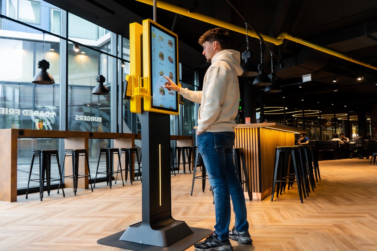 Gast bestellt an einem gelben Onesix Kiosk mit großem Touchscreen in einem Burgerrestaurant