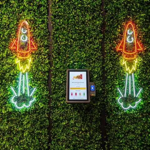 Onesix Kiosk Pro Wall an einer grünen Pflanzenwand zwischen Neon-Raketen