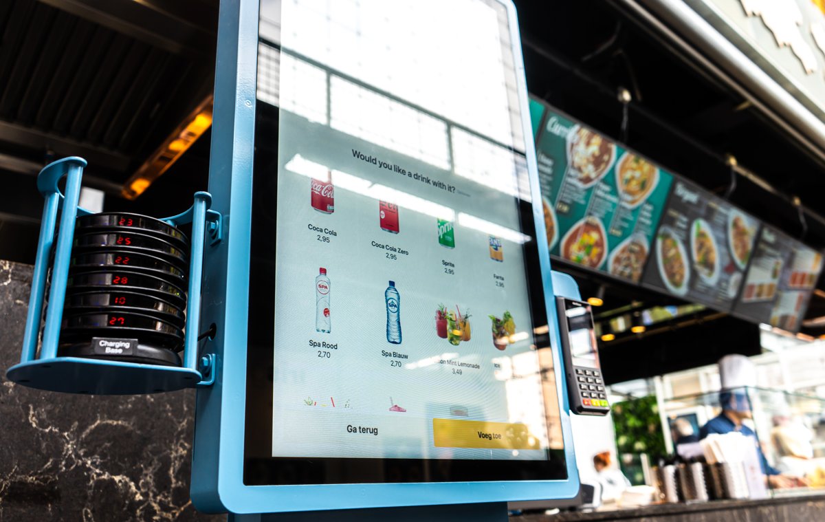 Onesix Kiosk Premium in einer belebten Markthalle, mit englischer Upsell-Frage auf dem Bestellbildschirm und Buzzern auf der Ladestation