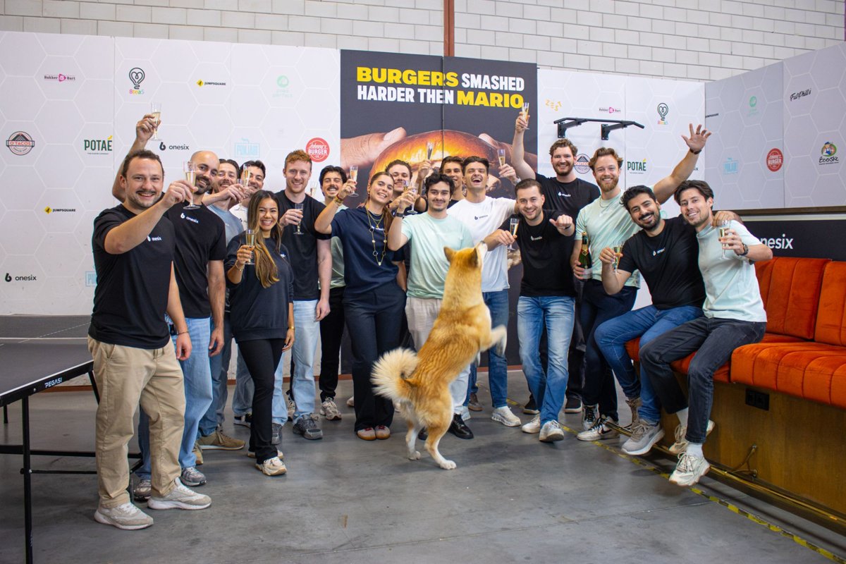 Das Onesix-Team stößt im Onesix-Büro mit Champagner an, im Vordergrund springt ein Hund