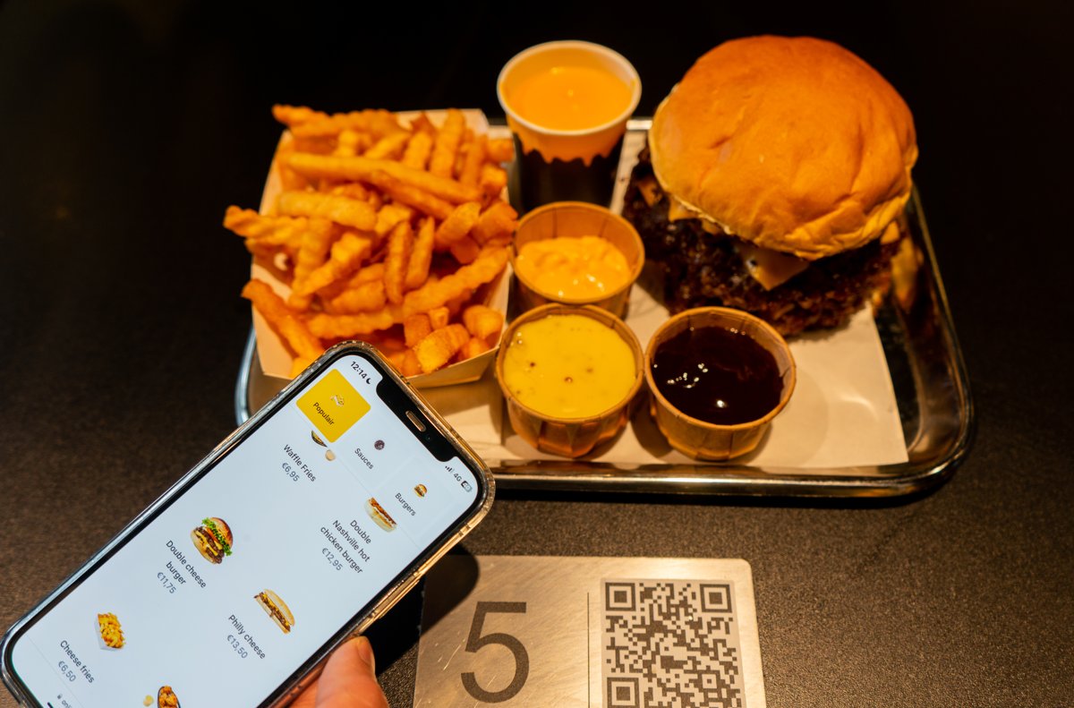 Gast schaut sich die digitale Speisekarte auf dem Handy an, neben einem Tischschild mit Tischnummer und QR-Code, dazu Burger und Pommes auf einem Tablett
