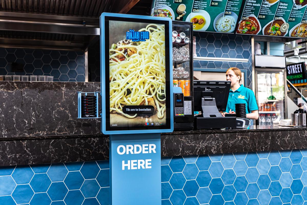 Blauer Onesix Kiosk mit „Order here“ neben der besetzten Theke in einem indischen Take-away-Restaurant